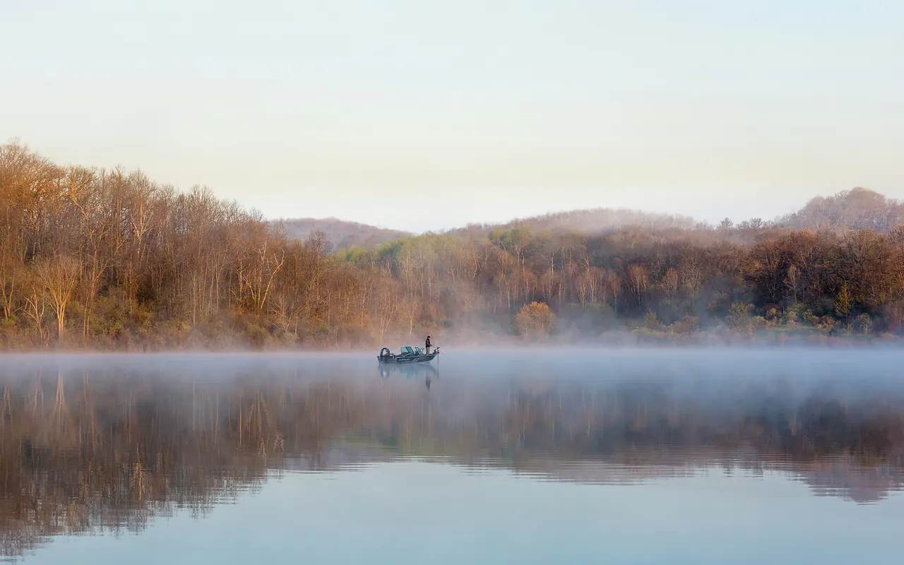 Nebliger See im Morgenlicht mit einem kleinen Boot und herbstlichem Ufer